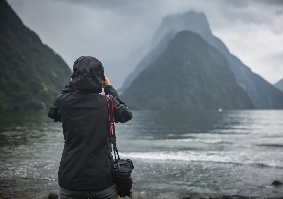 Visitor in a rain jacket photographing Mitre Peak in cloud at Milford Sound